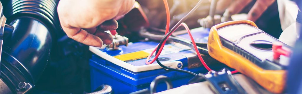 Close-up of a mechanic testing and connecting a car battery with diagnostic tools inside a vehicle engine bay.