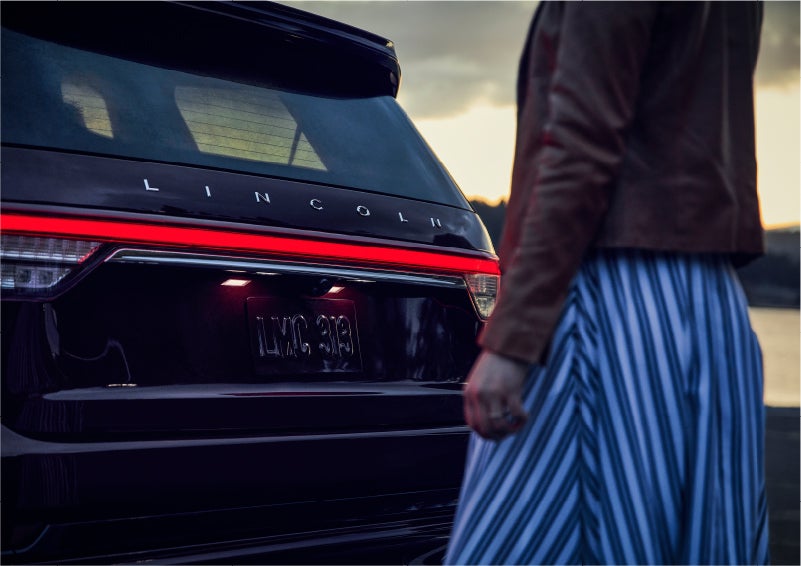 A person is shown near the rear of a 2023 Lincoln Aviator® SUV as the Lincoln Embrace illuminates the rear lights | West Point Lincoln of Sugar Land in Houston TX