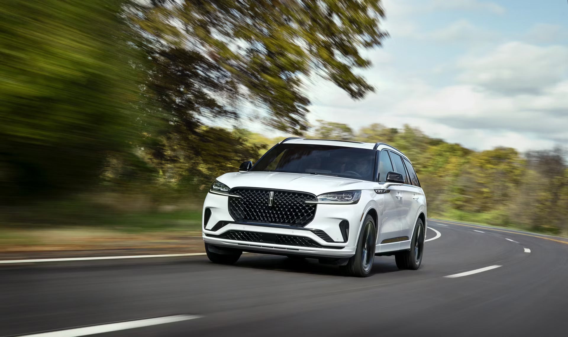White Lincoln Aviator SUV driving along a winding road surrounded by green trees.